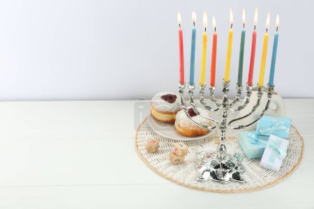Chanukka-Feier. Menora mit brennenden Kerzen, Dreidels, Donuts und Geschenkschachteln auf weißem Holztisch, Platz für Text