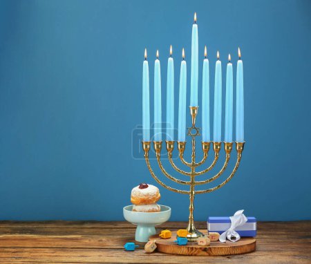 Chanukka-Feier. Menora mit brennenden Kerzen, Dreidels, Donuts und Geschenkbox auf Holztisch vor blauem Hintergrund, Platz für Text