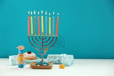 Hanoukka fête. Menorah avec des bougies allumées, des dreidels, des beignets et des boîtes-cadeaux sur une table en bois blanc sur fond bleu clair. Espace pour le texte