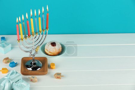 Chanukka-Feier. Menora mit brennenden Kerzen, Dreidels, Donut und Geschenkschachteln auf weißem Holztisch vor hellblauem Hintergrund. Raum für Text