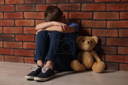 Kindesmissbrauch. Verärgerter Junge mit Teddybär sitzt auf dem Boden in der Nähe von Ziegelmauer