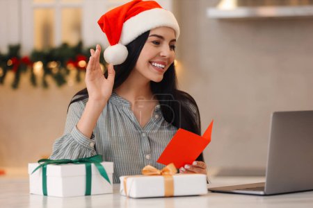 Celebrating Christmas online with exchanged by mail presents. Smiling woman in Santa hat with gifts waving hello during video call at home