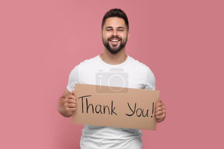 Happy man holding cardboard sheet with phrase Thank You on pink background
