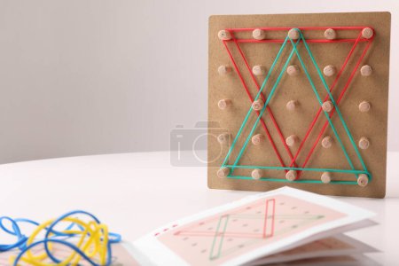 Holz-Geoboard mit Gummibändern und Anleitung auf weißem Tisch, Platz für Text. Motorische Entwicklung
