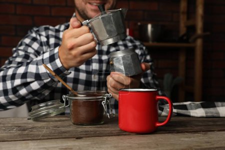 Man with moka pot in kitchen, focus on jar of ground coffee and mug on wooden table