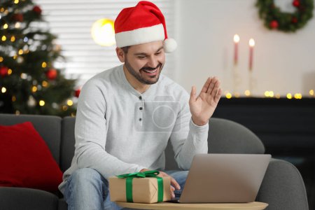 Celebrating Christmas online with exchanged by mail presents. Smiling man in Santa hat with gift waving hello during video call on laptop at home
