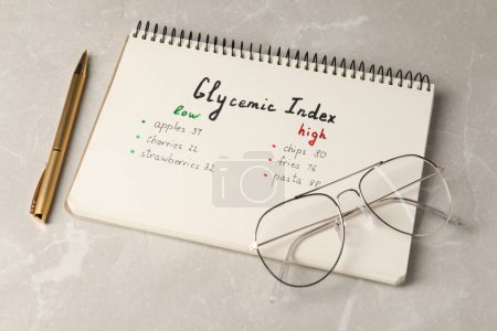 List with products of low and high glycemic index in notebook, pen and glasses on light grey marble table, above view