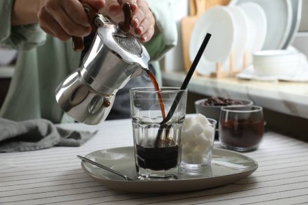 Woman pouring aromatic coffee from moka pot into glass at table in kitchen, closeup