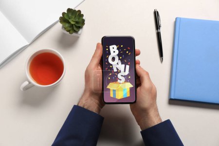 Bonus gaining. Man holding smartphone at white table, top view. Illustration of open gift box, word and confetti on device screen
