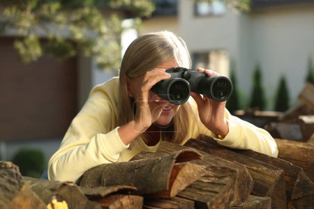Konzept des Privatlebens. Neugierige Seniorin mit Fernglas spioniert Nachbarn über Brennholz im Freien aus