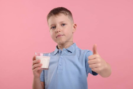 Téléchargez les photos : Mignon garçon avec un verre de lait frais montrant pouce vers le haut sur fond rose - en image libre de droit