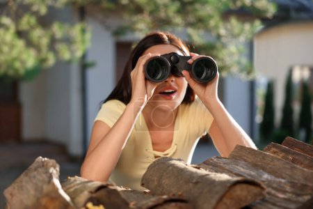 Konzept des Privatlebens. Neugierige junge Frau mit Fernglas spioniert Nachbarn über Brennholz im Freien aus