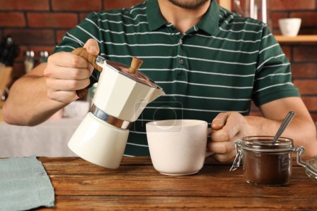 Mann schüttet aromatischen Kaffee aus Mokka-Kanne in Tasse an Holztisch drinnen, Nahaufnahme