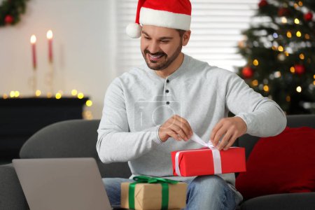 Célébrons Noël en ligne avec des cadeaux échangés par la poste. Homme heureux dans la boîte cadeau d'ouverture de chapeau de père Noël pendant l'appel vidéo sur l'ordinateur portable à la maison