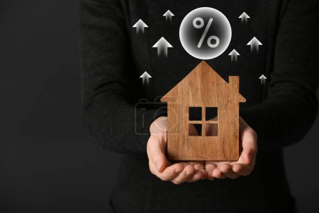 Steigende Hypothekenzinsen illustriert durch Aufwärtspfeile und Prozentzeichen. Frau mit Holzhaus auf dunklem Hintergrund, Nahaufnahme