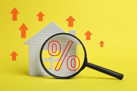 Hausse du taux hypothécaire illustrée par des flèches vers le haut et un signe en pourcentage. Modèle de maison et loupe sur fond jaune