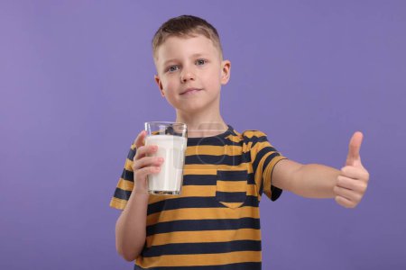 Téléchargez les photos : Mignon garçon avec un verre de lait frais montrant pouce vers le haut sur fond violet - en image libre de droit