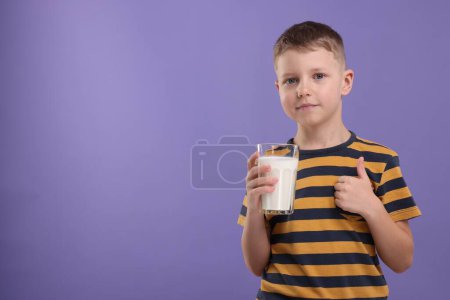 Téléchargez les photos : Garçon mignon avec un verre de lait frais montrant pouce vers le haut sur fond violet, espace pour le texte - en image libre de droit
