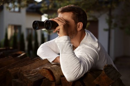 Konzept des Privatlebens. Neugieriger Mann mit Fernglas spioniert Nachbarn über Brennholz im Freien aus