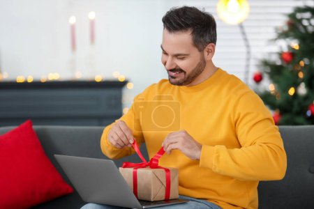 Weihnachten online feiern und Geschenke per Post austauschen. Glücklicher Mann öffnet Geschenkbox während Videoanruf auf Laptop zu Hause