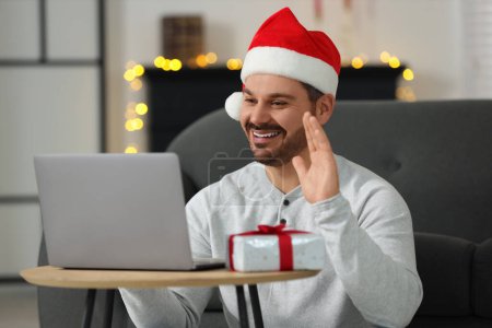 Celebrating Christmas online with exchanged by mail presents. Smiling man in Santa hat with gift waving hello during video call on laptop at home