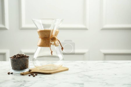 Empty glass chemex coffeemaker and beans in bowl on white marble table, space for text