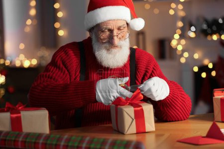 Foto de Santa Claus atando arco en caja de regalo en su lugar de trabajo en la habitación decorada para Navidad - Imagen libre de derechos