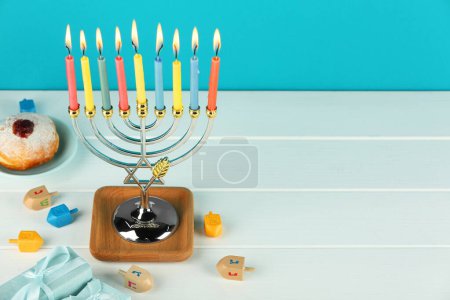 Hanukkah celebration. Menorah with burning candles, dreidels, donut and gift boxes on white wooden table against light blue background. Space for text-stock-photo