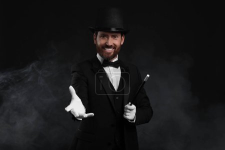 Happy magician holding wand in smoke on black background