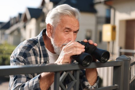 Konzept des Privatlebens. Neugieriger Senior mit Fernglas spioniert Nachbarn über Zaun im Freien aus