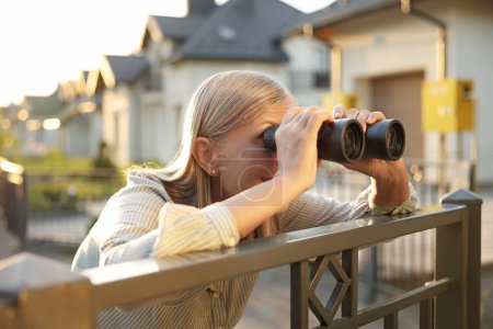 Photo for Concept of private life. Curious senior woman with binoculars spying on neighbours over fence outdoors - Royalty Free Image
