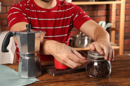 Kaffee brauen. Mann mit Glas Bohnen, Mokka-Topf und Becher an Holztisch drinnen, Nahaufnahme
