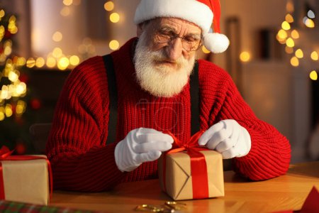 Foto de Santa Claus atando arco en caja de regalo en su lugar de trabajo en la habitación decorada para Navidad - Imagen libre de derechos