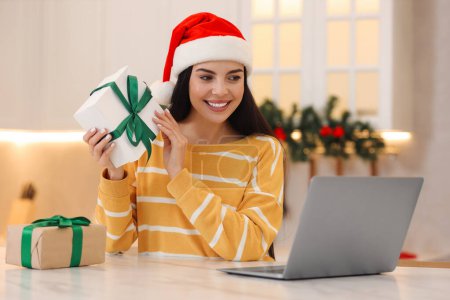 Celebrando la Navidad en línea con regalos intercambiados por correo. Mujer sonriente en Santa sombrero con cajas de regalo durante la videollamada en el ordenador portátil en casa