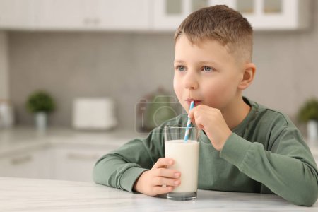 Téléchargez les photos : Mignon garçon boire du lait frais à partir de verre à la table blanche dans la cuisine, espace pour le texte - en image libre de droit