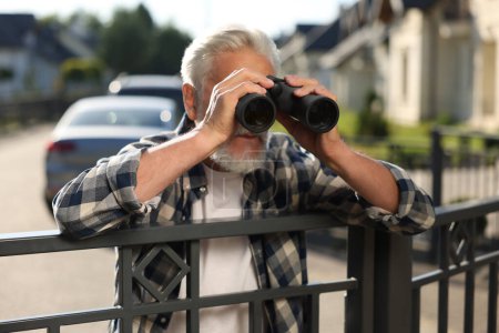 Téléchargez les photos : Concept de vie privée. Homme âgé curieux avec des jumelles espionnant les voisins au-dessus de la clôture à l'extérieur - en image libre de droit