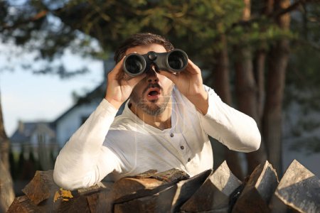 Téléchargez les photos : Concept de vie privée. Homme curieux avec des jumelles espionnant les voisins sur du bois de chauffage à l'extérieur - en image libre de droit