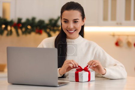 Célébrons Noël en ligne avec des cadeaux échangés par la poste. Souriant femme ouverture boîte cadeau pendant appel vidéo sur ordinateur portable à la maison