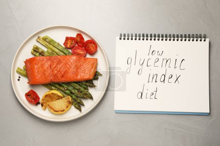 Notebook with words Low Glycemic Index Diet and plate of tasty grilled salmon on grey table, flat lay