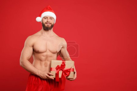 Photo for Attractive young man with muscular body in Santa hat holding Christmas gift box on red background, space for text - Royalty Free Image