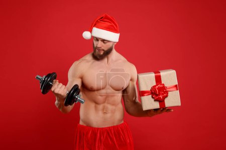 Photo for Muscular young man in Santa hat with dumbbell and Christmas gift box on red background - Royalty Free Image