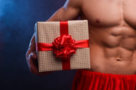 Photo for Young man with muscular body holding Christmas gift box on color background, closeup - Royalty Free Image