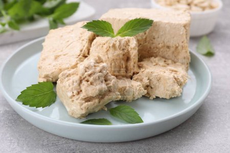 Plate with pieces of tasty halva and mint leaves on light grey table, closeup