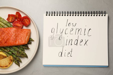 Cuaderno con palabras Bajo índice glucémico Dieta y plato de sabrosa mesa gris salmón a la parrilla, puesta plana
