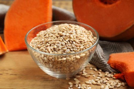 Dry pearl barley in bowl and pieces of pumpkin on wooden table, closeup