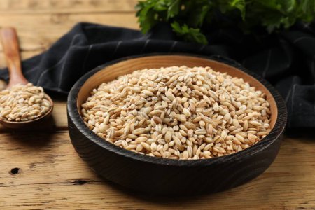 Dry pearl barley in bowl and spoon on wooden table, closeup