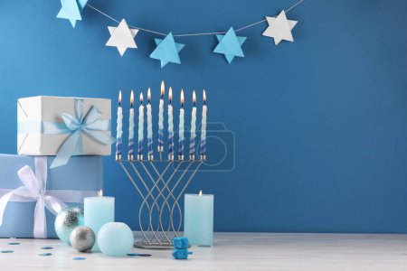 Chanukka-Feier. Menora, brennende Kerzen, Dreidel und Geschenkschachteln auf weißem Holztisch vor hellblauem Hintergrund, Platz für Text