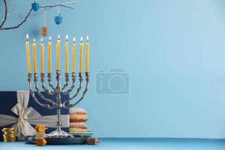 Hanukkah celebration. Menorah with burning candles, dreidels, donuts and gift box on table against light blue background. Space for text