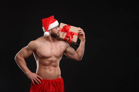 Photo for Attractive young man with muscular body holding Christmas gift box on black background, space for text - Royalty Free Image