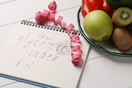 Cuaderno con palabras Índice glucémico bajo Dieta, cinta métrica y productos saludables en mesa de madera blanca, primer plano